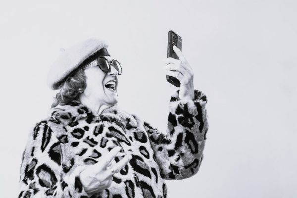 Stylish senior woman in animal print coat, sunglasses and beret using her mobile phone while standing on an yellow background. Technology concept.